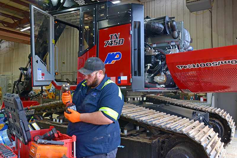 A heavy equipment mechanic in safety goggles and gloves holding an impact driver while a large excavator stands in the background with its engine compartment open